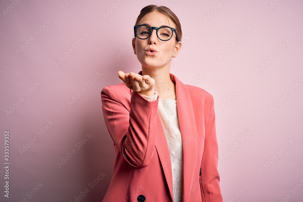 Young beautiful redhead woman wearing jacket and glasses over isolated pink background looking at the camera blowing a kiss with hand on air being lovely and sexy. Love expression.
