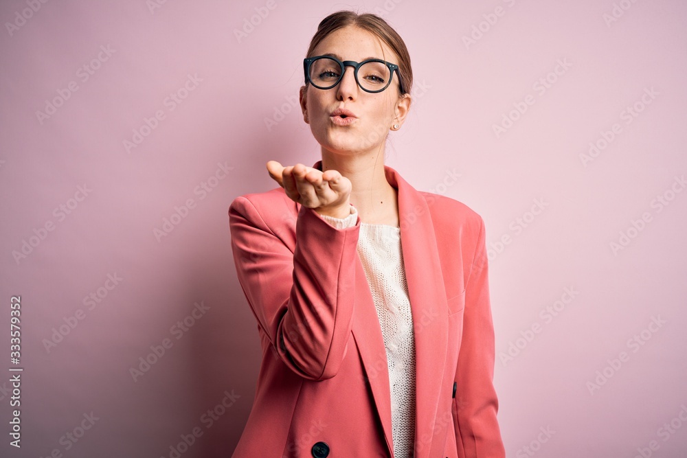Young beautiful redhead woman wearing jacket and glasses over isolated pink background looking at the camera blowing a kiss with hand on air being lovely and sexy. Love expression. Young beautiful redhead woman wearing jacket and glasses over isolated pink background looking at the camera blowing a kiss with hand on air being lovely and sexy. Love expression.
