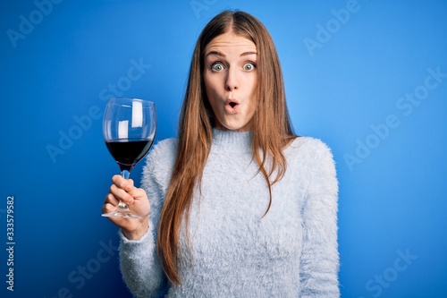 Photographie Young beautiful redhead woman drinking glass of red wine over isolated blue back