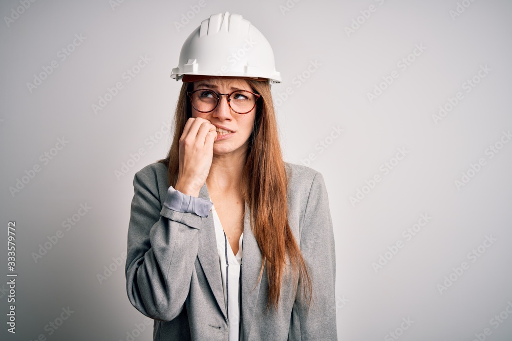 Young beautiful redhead architect woman wearing security helmet over white background looking stressed and nervous with hands on mouth biting nails. Anxiety problem.