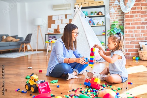 Caucasian girl kid playing and learning at playschool with female teacher. Mother and daughter at playroom around toys playing with bulding blocks