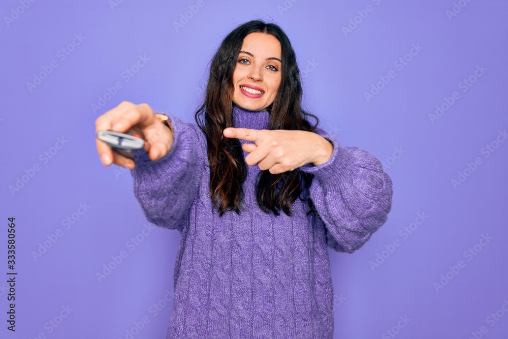 Young beautiful woman using television remote control over isolated purple background very happy pointing with hand and finger Young beautiful woman using television remote control over isolated purple background very happy pointing with hand and finger