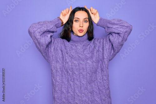 Young beautiful woman wearing casual turtleneck sweater standing over purple background doing funny gesture with finger over head as bull horns © Krakenimages.com