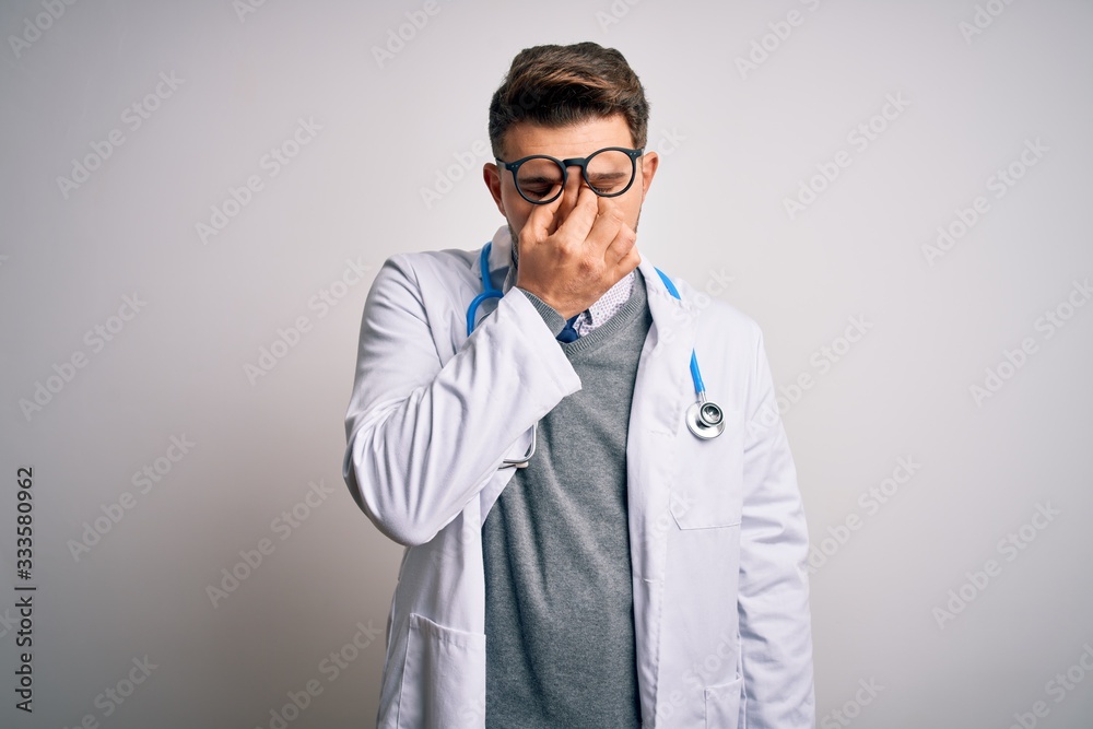 Young doctor man with blue eyes wearing medical coat and stethoscope over isolated background tired rubbing nose and eyes feeling fatigue and headache. Stress and frustration concept. Young doctor man with blue eyes wearing medical coat and stethoscope over isolated background tired rubbing nose and eyes feeling fatigue and headache. Stress and frustration concept.
