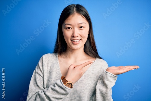 Young beautiful asian woman wearing casual sweater standing over blue isolated background amazed and smiling to the camera while presenting with hand and pointing with finger.
