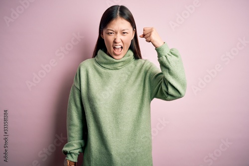 Young beautiful asian woman wearing green winter sweater over pink solated background angry and mad raising fist frustrated and furious while shouting with anger. Rage and aggressive concept. © Krakenimages.com