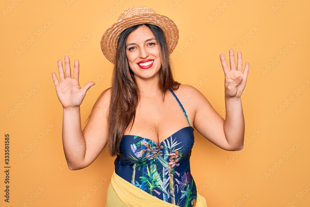 Young hispanic woman wearing summer hat and swimsuit over yellow background showing and pointing up with fingers number nine while smiling confident and happy. Young hispanic woman wearing summer hat and swimsuit over yellow background showing and pointing up with fingers number nine while smiling confident and happy.