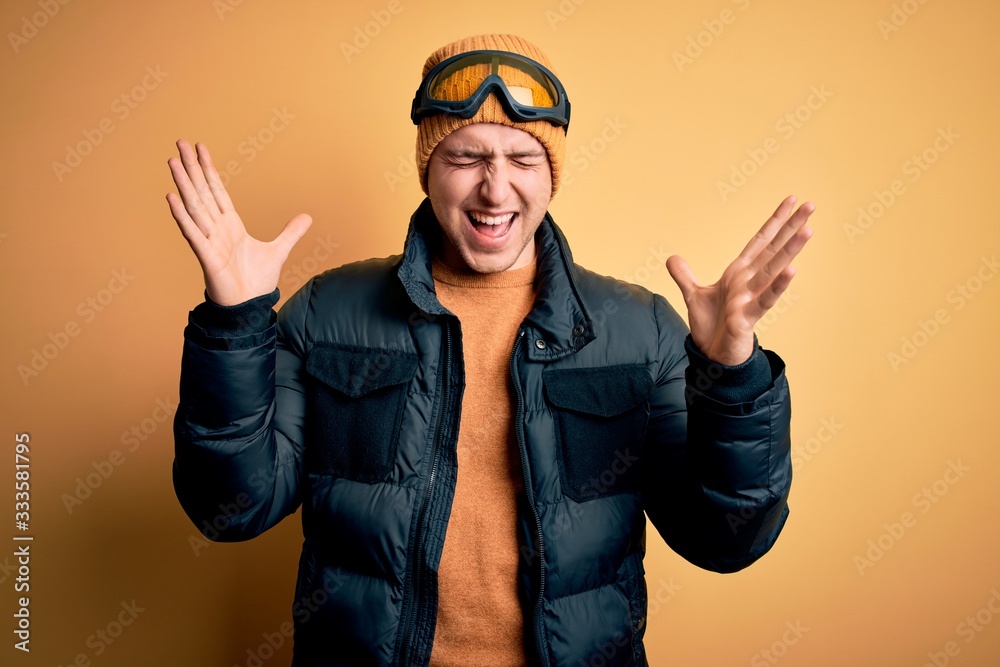 Young handsome caucasian man wearing hat, coat and ski glasses for winter and snow weather celebrating mad and crazy for success with arms raised and closed eyes screaming excited. Winner concept Young handsome caucasian man wearing hat, coat and ski glasses for winter and snow weather celebrating mad and crazy for success with arms raised and closed eyes screaming excited. Winner concept