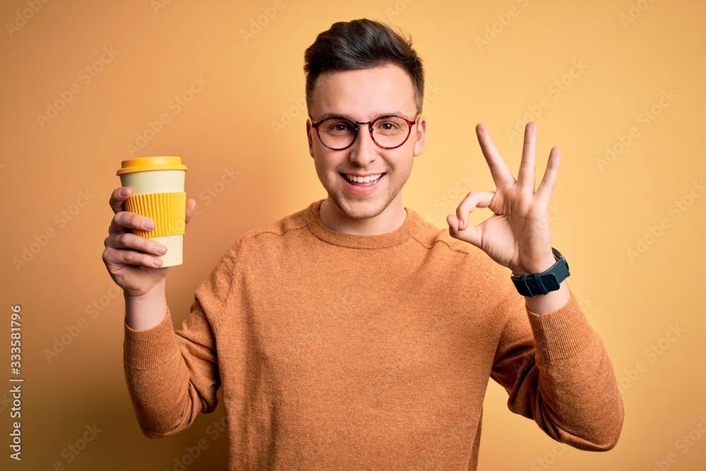 Young handsome caucasian man drinking coffee from a take away thermo bottle doing ok sign with fingers, excellent symbol Young handsome caucasian man drinking coffee from a take away thermo bottle doing ok sign with fingers, excellent symbol
