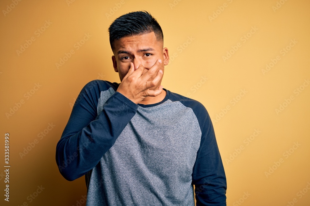 Young handsome latin man wearing casual t-shirt standing over yellow background smelling something stinky and disgusting, intolerable smell, holding breath with fingers on nose. Bad smell
