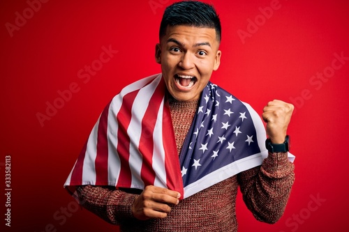 Young handsome latin patriotic man wearing united states of america falg screaming proud and celebrating victory and success very excited, cheering emotion photo