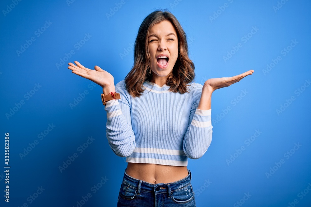 Young beautiful blonde girl wearing casual sweater standing over blue isolated background celebrating mad and crazy for success with arms raised and closed eyes screaming excited. Winner concept