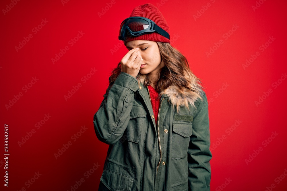 Young blonde girl wearing ski glasses and winter coat for ski weather over red background tired rubbing nose and eyes feeling fatigue and headache. Stress and frustration concept. Young blonde girl wearing ski glasses and winter coat for ski weather over red background tired rubbing nose and eyes feeling fatigue and headache. Stress and frustration concept.