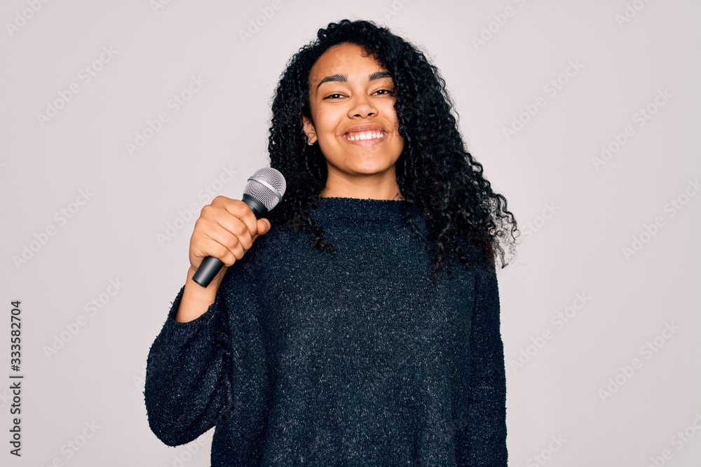 Young african american curly singer woman singing using microphone over white background with a happy face standing and smiling with a confident smile showing teeth Young african american curly singer woman singing using microphone over white background with a happy face standing and smiling with a confident smile showing teeth