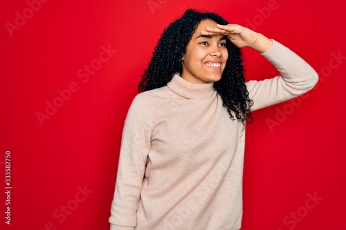 Young african american curly woman wearing casual turtleneck sweater over red background very happy and smiling looking far away with hand over head. Searching concept. © Krakenimages.com