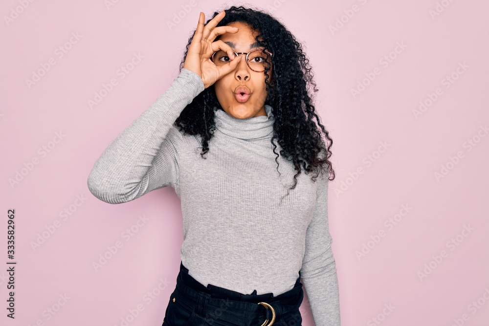Young african american woman wearing turtleneck sweater and glasses over pink background doing ok gesture shocked with surprised face, eye looking through fingers. Unbelieving expression. Young african american woman wearing turtleneck sweater and glasses over pink background doing ok gesture shocked with surprised face, eye looking through fingers. Unbelieving expression.