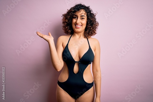 Young beautiful arab woman on vacation wearing swimsuit and sunglasses over pink background smiling cheerful presenting and pointing with palm of hand looking at the camera.