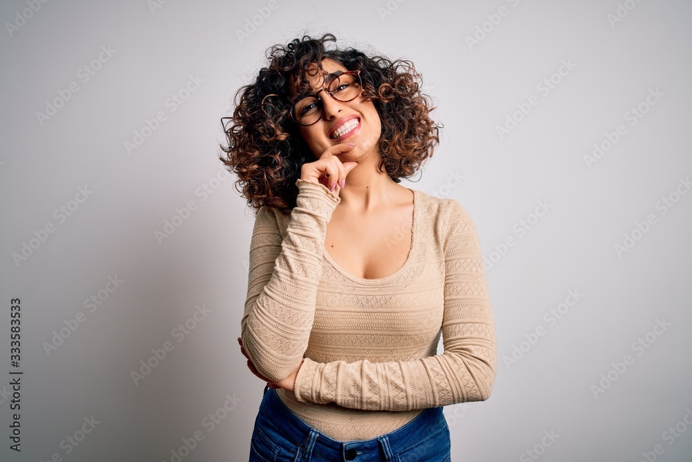 Young beautiful curly arab woman wearing casual t-shirt and glasses over white background looking confident at the camera smiling with crossed arms and hand raised on chin. Thinking positive. Young beautiful curly arab woman wearing casual t-shirt and glasses over white background looking confident at the camera smiling with crossed arms and hand raised on chin. Thinking positive.