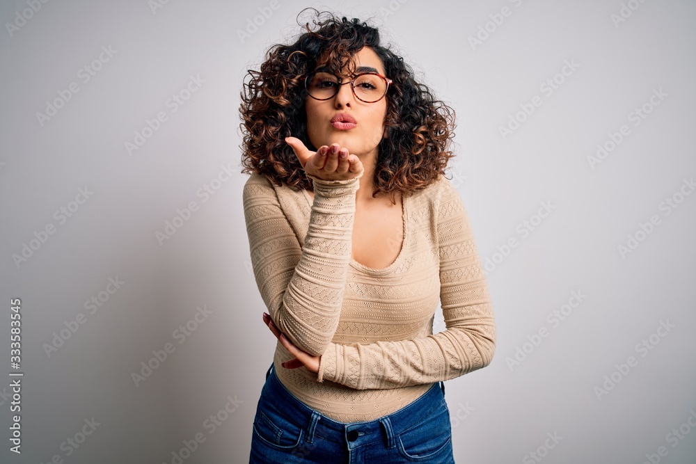 Young beautiful curly arab woman wearing casual t-shirt and glasses over white background looking at the camera blowing a kiss with hand on air being lovely and sexy. Love expression. Young beautiful curly arab woman wearing casual t-shirt and glasses over white background looking at the camera blowing a kiss with hand on air being lovely and sexy. Love expression.