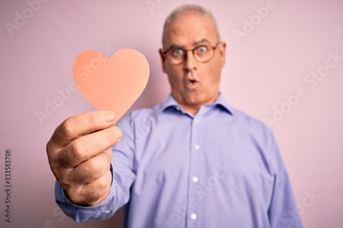 Middle age hoary romantic man holding red paper heart shape over isolated pink background scared in shock with a surprise face, afraid and excited with fear expression