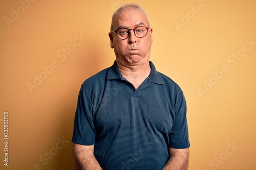 Middle age handsome hoary man wearing casual polo and glasses over yellow background puffing cheeks with funny face. Mouth inflated with air, crazy expression.