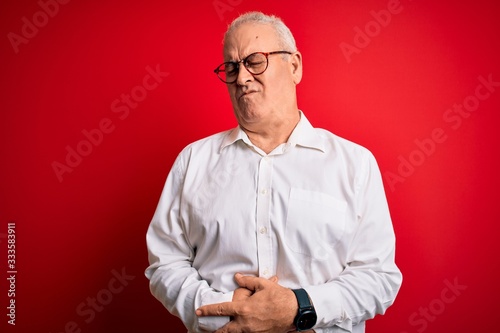 Middle age handsome hoary man wearing casual shirt and glasses over red background with hand on stomach because indigestion, painful illness feeling unwell. Ache concept.