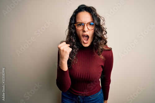 Beautiful woman with curly hair wearing casual sweater and glasses over white background angry and mad raising fist frustrated and furious while shouting with anger. Rage and aggressive concept.
