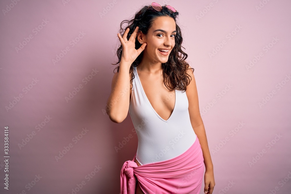 Beautiful woman with curly hair on vacation wearing white swimsuit over pink background smiling with hand over ear listening an hearing to rumor or gossip. Deafness concept. Beautiful woman with curly hair on vacation wearing white swimsuit over pink background smiling with hand over ear listening an hearing to rumor or gossip. Deafness concept.