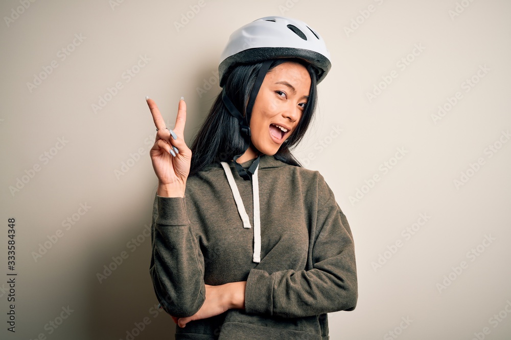Young beautiful chinese woman wearing bike helmet over isolated white background smiling with happy face winking at the camera doing victory sign. Number two. Young beautiful chinese woman wearing bike helmet over isolated white background smiling with happy face winking at the camera doing victory sign. Number two.