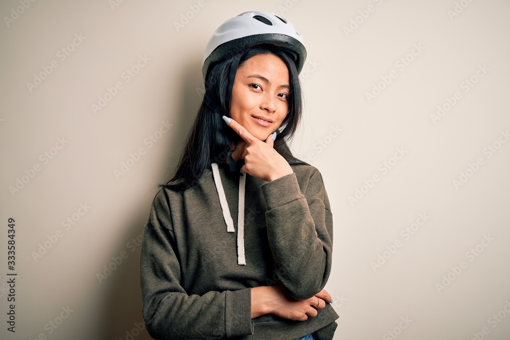 Young beautiful chinese woman wearing bike helmet over isolated white background looking confident at the camera smiling with crossed arms and hand raised on chin. Thinking positive. Young beautiful chinese woman wearing bike helmet over isolated white background looking confident at the camera smiling with crossed arms and hand raised on chin. Thinking positive.