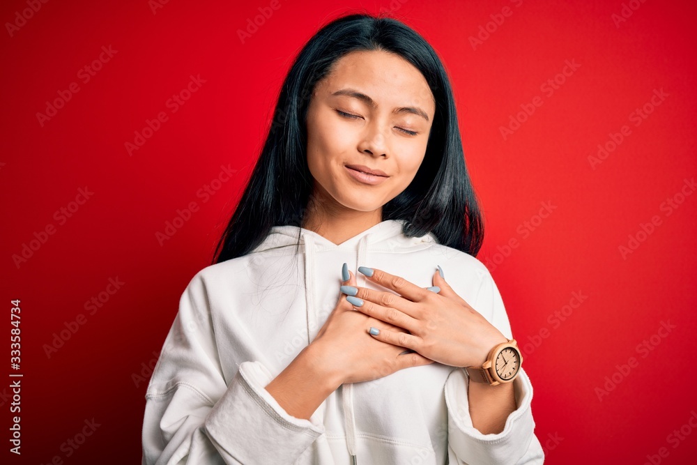 Young beautiful chinese sporty woman wearing sweatshirt over isolated red background smiling with hands on chest with closed eyes and grateful gesture on face. Health concept.