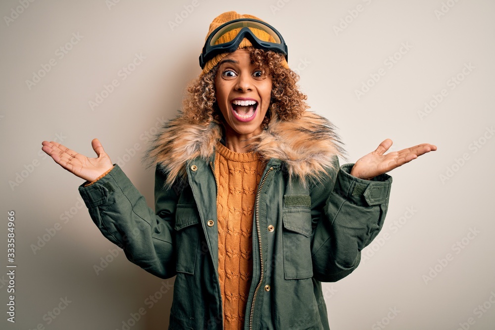 Young african american skier woman with curly hair wearing snow sportswear and ski goggles celebrating crazy and amazed for success with arms raised and open eyes screaming excited. Winner concept Young african american skier woman with curly hair wearing snow sportswear and ski goggles celebrating crazy and amazed for success with arms raised and open eyes screaming excited. Winner concept