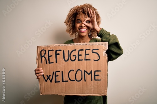 African american woman asking for immigration holding banner with wlecome refugees message with happy face smiling doing ok sign with hand on eye looking through fingers