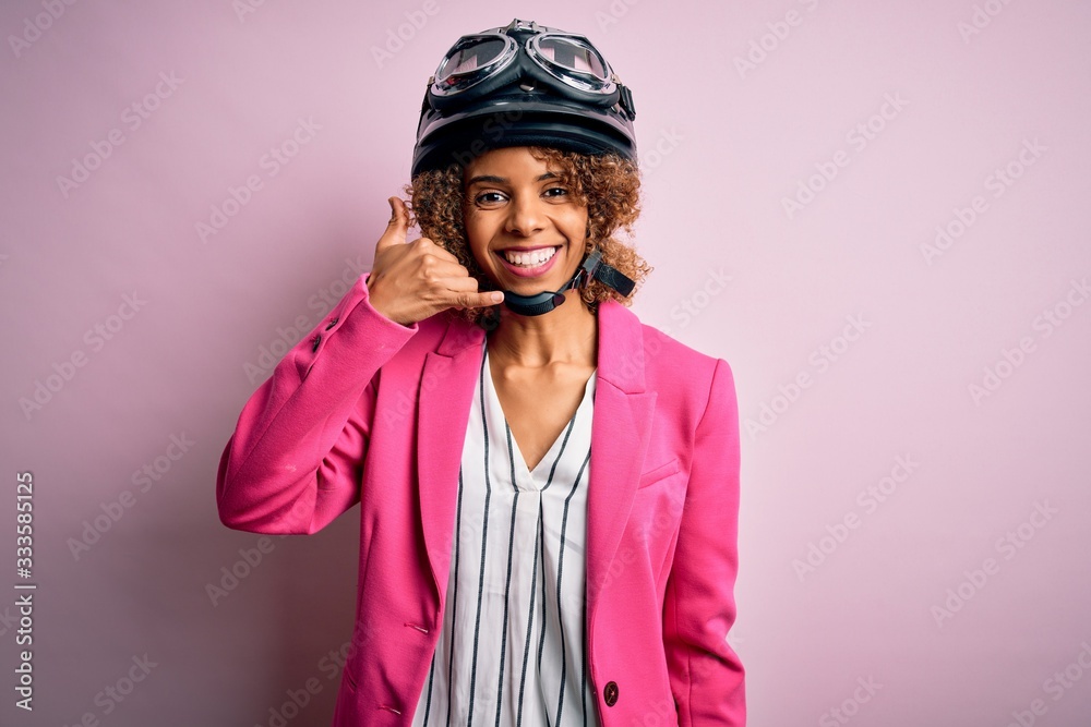 African american motorcyclist woman with curly hair wearing moto helmet over pink background smiling doing phone gesture with hand and fingers like talking on the telephone. Communicating concepts.