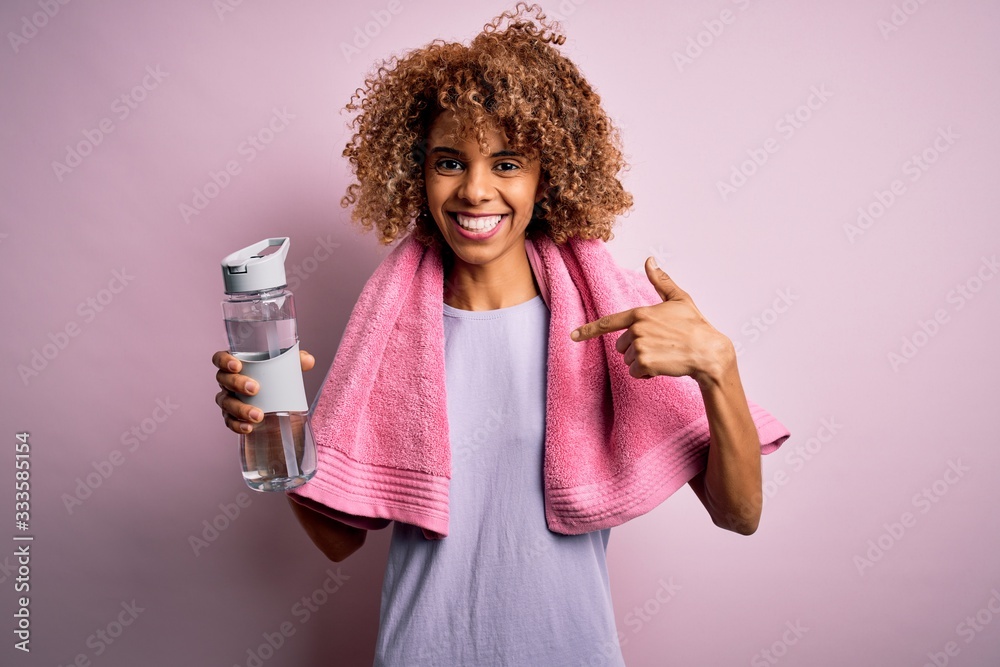 Young african american sportswoman doing sport wearing towel drinking bottle of water with surprise face pointing finger to himself Young african american sportswoman doing sport wearing towel drinking bottle of water with surprise face pointing finger to himself