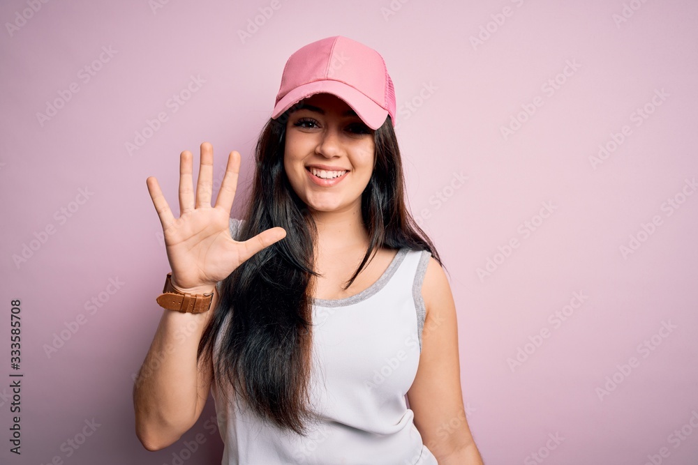 Young brunette woman wearing casual sport cap over pink background showing and pointing up with fingers number five while smiling confident and happy. Young brunette woman wearing casual sport cap over pink background showing and pointing up with fingers number five while smiling confident and happy.