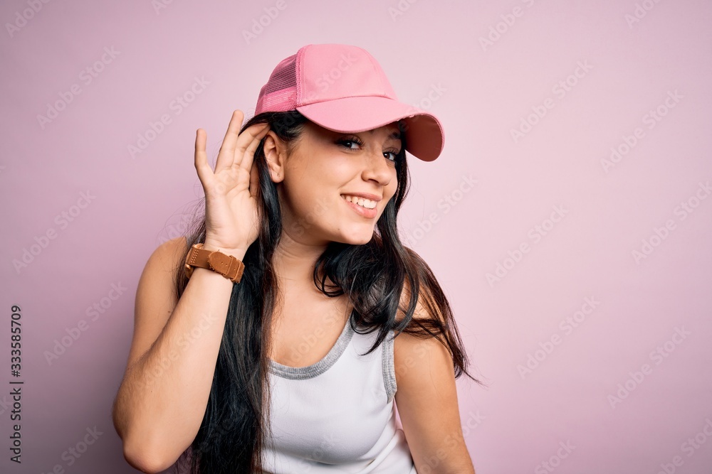 Young brunette woman wearing casual sport cap over pink background smiling with hand over ear listening an hearing to rumor or gossip. Deafness concept. Young brunette woman wearing casual sport cap over pink background smiling with hand over ear listening an hearing to rumor or gossip. Deafness concept.