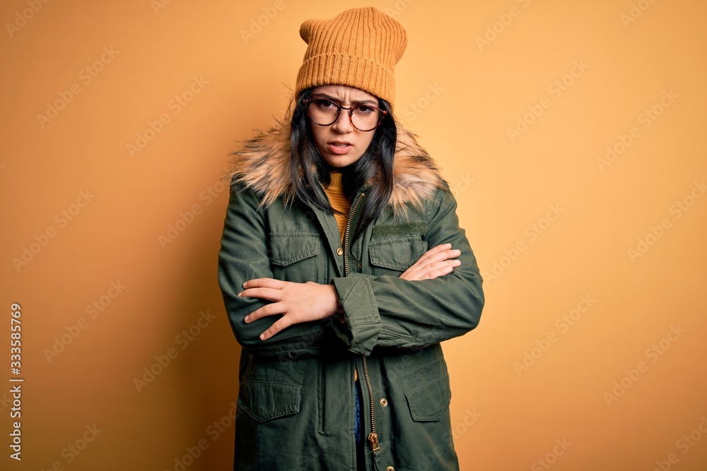 Young brunette woman wearing glasses and winter coat with hat over yellow isolated background skeptic and nervous, disapproving expression on face with crossed arms. Negative person.