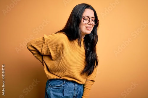 Young brunette woman wearing glasses and casual sweater over yellow isolated background Suffering of backache, touching back with hand, muscular pain