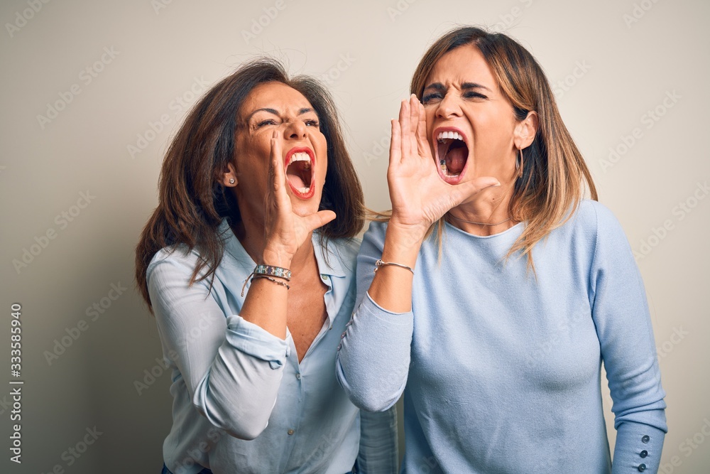 Middle age beautiful couple of sisters standing over isolated white background shouting and screaming loud to side with hand on mouth. Communication concept. Middle age beautiful couple of sisters standing over isolated white background shouting and screaming loud to side with hand on mouth. Communication concept.