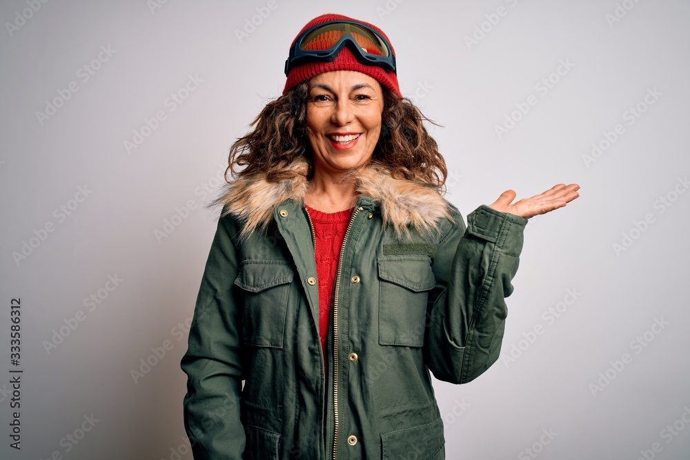 Middle age skier woman wearing snow sportswear and ski goggles over white background smiling cheerful presenting and pointing with palm of hand looking at the camera.