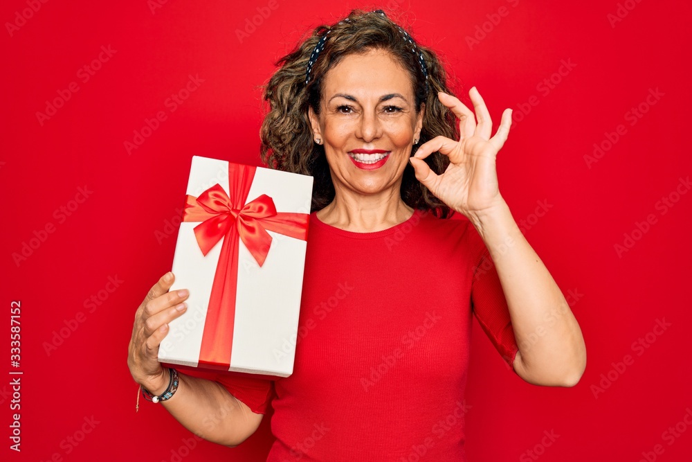 Middle age senior woman holding romantic present box over red isolated background doing ok sign with fingers, excellent symbol Middle age senior woman holding romantic present box over red isolated background doing ok sign with fingers, excellent symbol
