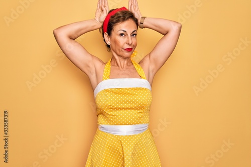Middle age senior pin up woman wearing 50s style retro dress over yellow background Doing bunny ears gesture with hands palms looking cynical and skeptical. Easter rabbit concept.