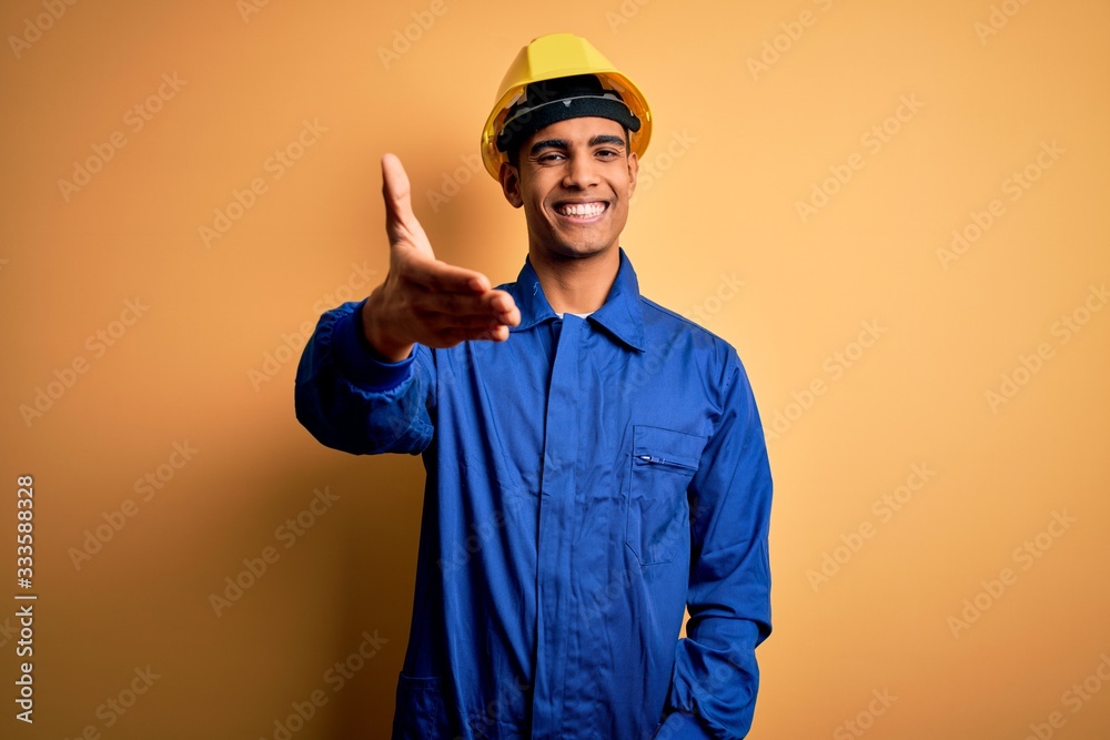 Young handsome african american worker man wearing blue uniform and security helmet smiling friendly offering handshake as greeting and welcoming. Successful business.