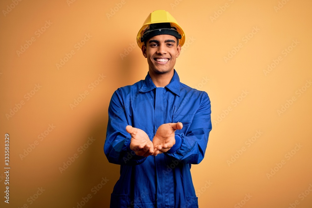 Young handsome african american worker man wearing blue uniform and security helmet Smiling with hands palms together receiving or giving gesture. Hold and protection