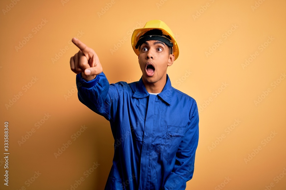 Young handsome african american worker man wearing blue uniform and security helmet Pointing with finger surprised ahead, open mouth amazed expression, something on the front