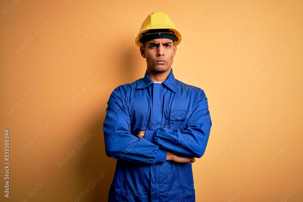 Young handsome african american worker man wearing blue uniform and security helmet skeptic and nervous, disapproving expression on face with crossed arms. Negative person.
