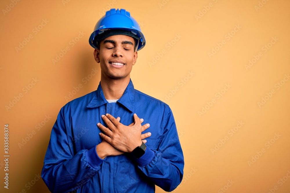 Young handsome african american worker man wearing blue uniform and security helmet smiling with hands on chest with closed eyes and grateful gesture on face. Health concept.
