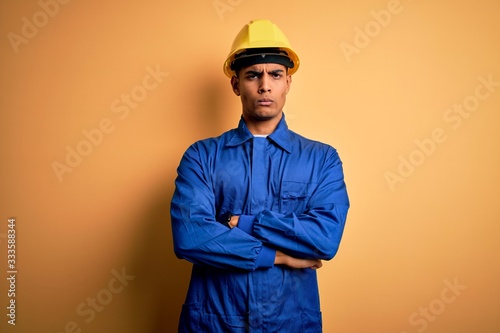 Young handsome african american worker man wearing blue uniform and security helmet skeptic and nervous, disapproving expression on face with crossed arms. Negative person.