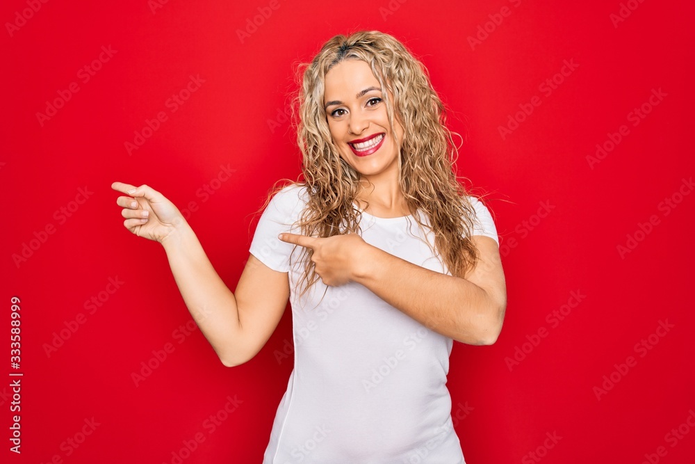 Young beautiful blonde woman wearing casual t-shirt standing over isolated red background smiling and looking at the camera pointing with two hands and fingers to the side. Young beautiful blonde woman wearing casual t-shirt standing over isolated red background smiling and looking at the camera pointing with two hands and fingers to the side.